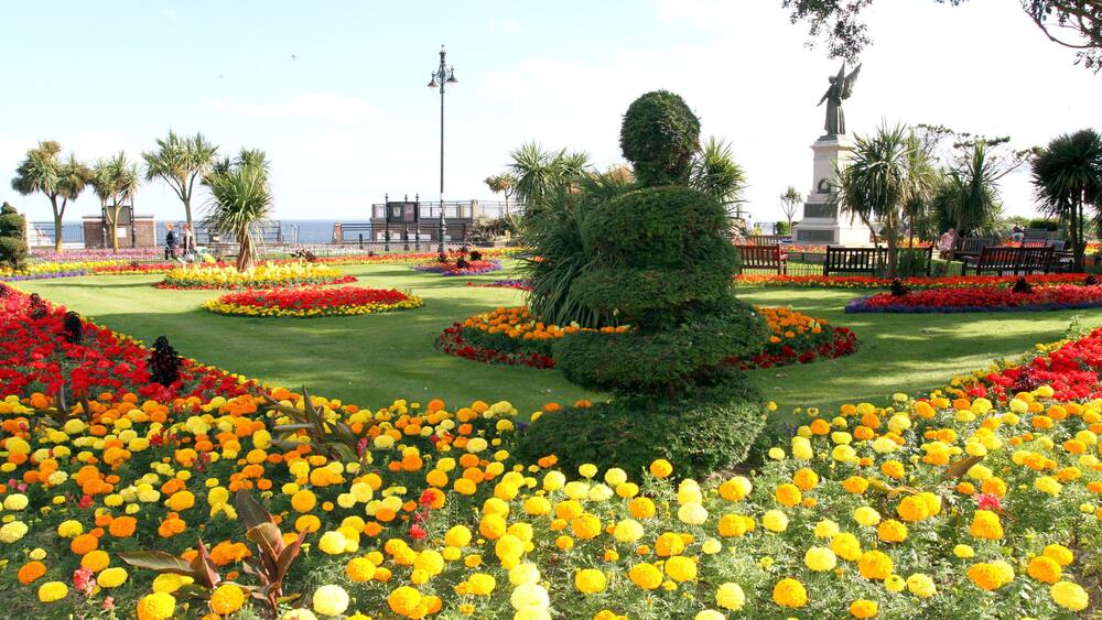 A landscaped garden with vibrant flower beds, a central topiary, and a statue.