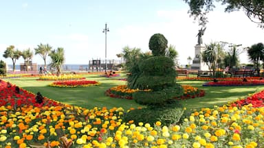 Um jardim paisagístico com canteiros de flores vibrantes, uma topiária central e uma estátua.