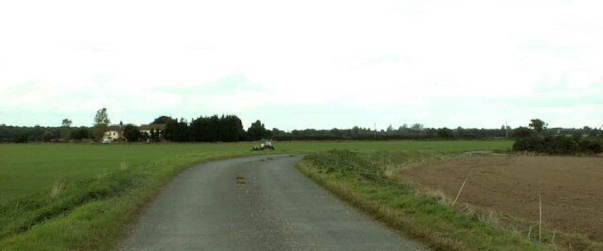 A rural road curving through fields with a farmhouse in the distance.