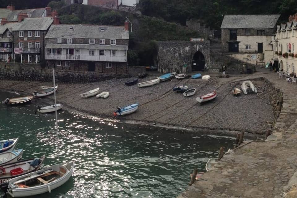 A coastal village with boats docked along the shore, a stone wall, and houses on a hillside.