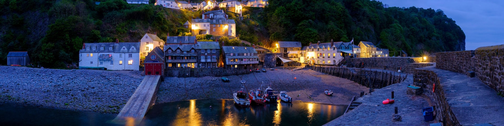 A coastal village at dusk with illuminated buildings, a calm harbor, and a stone wall.