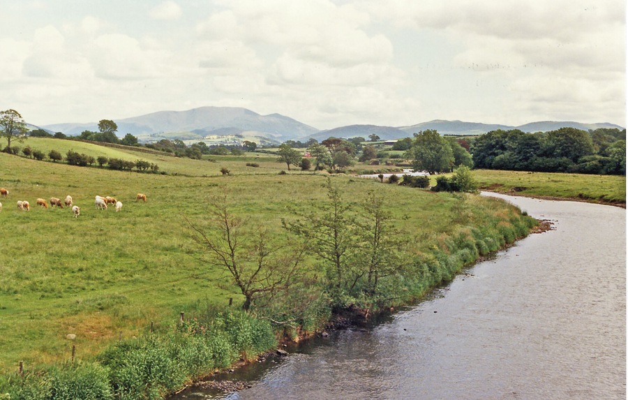 Upstream on River Derwent at Great Broughton, 1986. View eastward from the bridge between Great Broughton (to left) and the A66 (to right). On the horizon is Skiddaw (3,054 ft.) and nearer to its right the fells over the west shore of Bassenthwaite Lake.