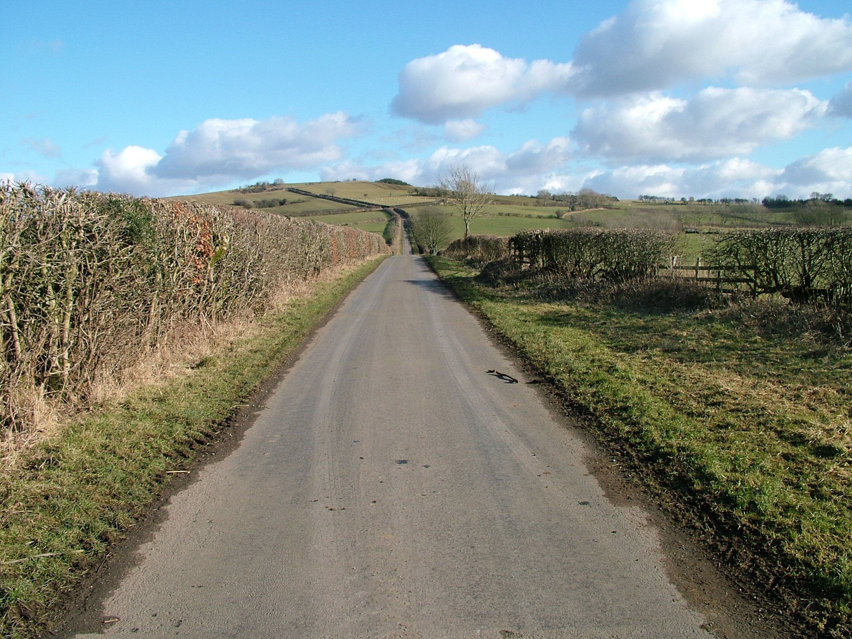 A rural road flanked by fields and stone walls under a partly cloudy sky.