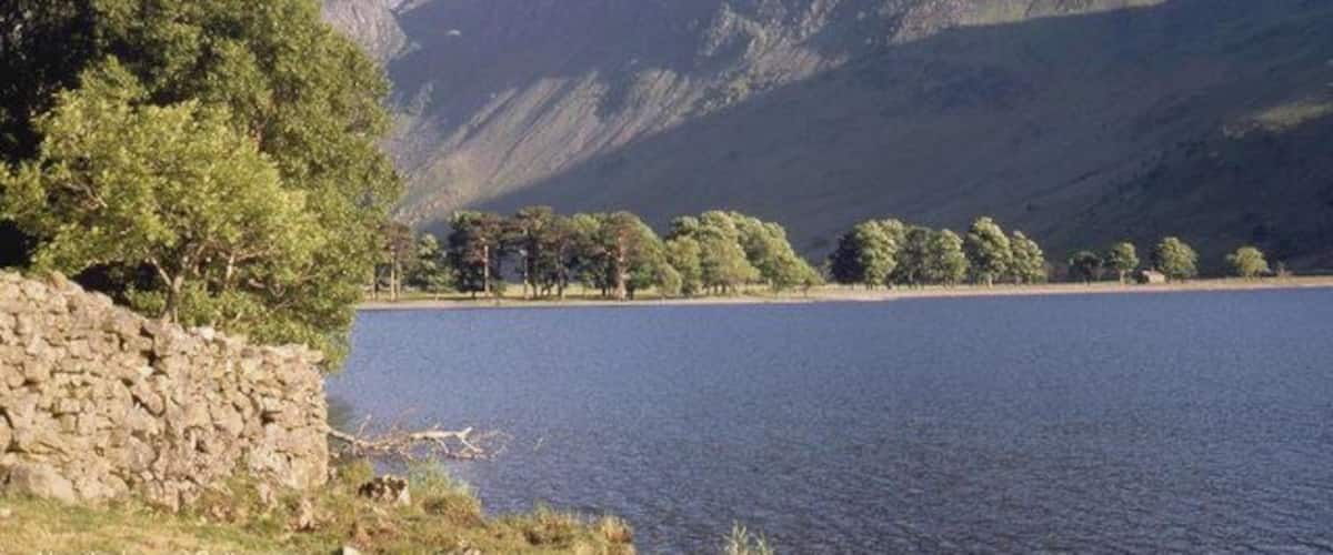A lake with a stone wall on the left, surrounded by trees and mountains in the background.
