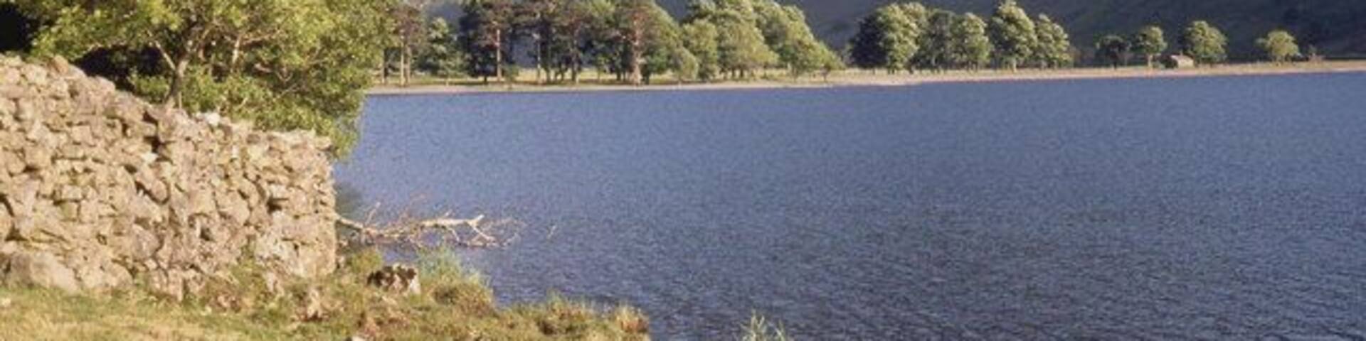 A lake with a stone wall on the left, surrounded by trees and mountains in the background.