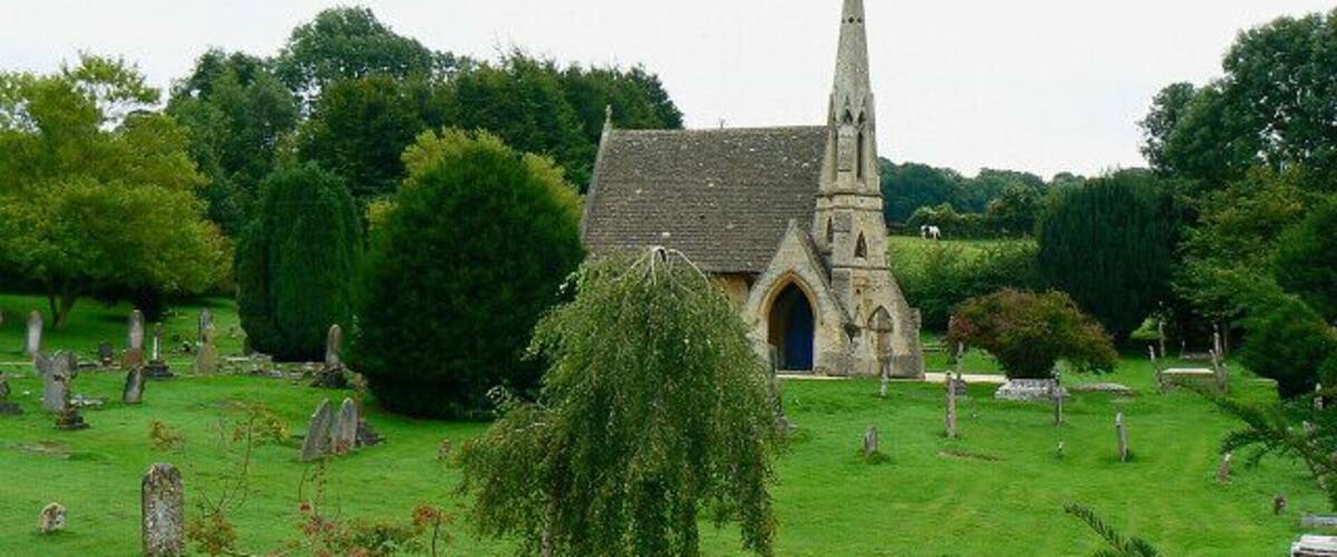 A church with a tall spire, surrounded by a cemetery with gravestones and lush greenery.