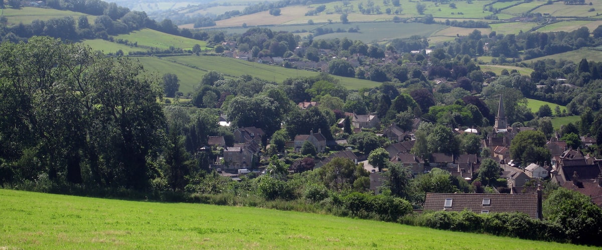 A rural landscape with rolling hills, scattered houses, and dense tree coverage.