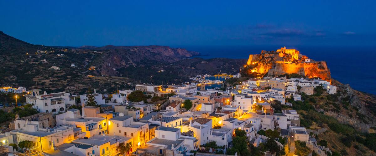 A coastal town at dusk with illuminated buildings and a prominent fortified hill in the background.