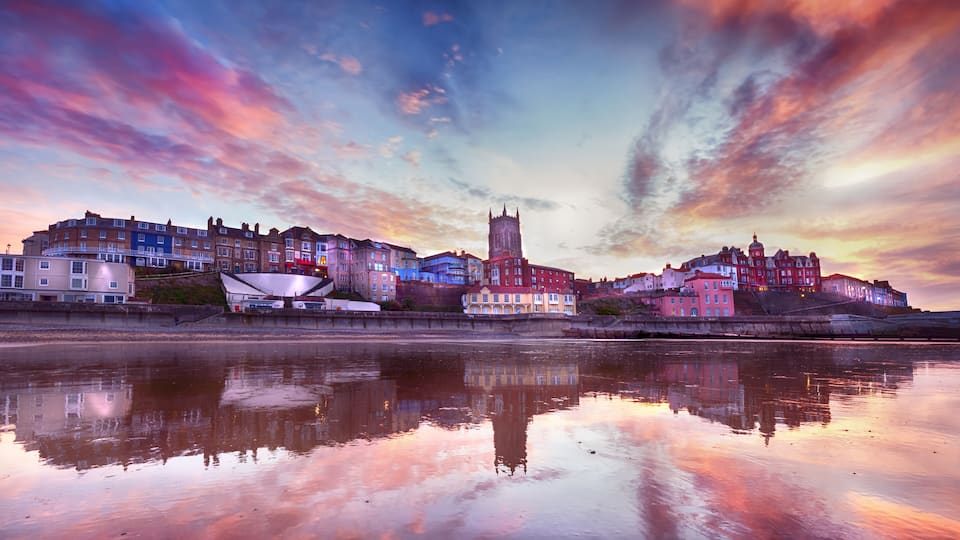 A waterfront cityscape with historic buildings and a prominent tower reflecting in the calm water.