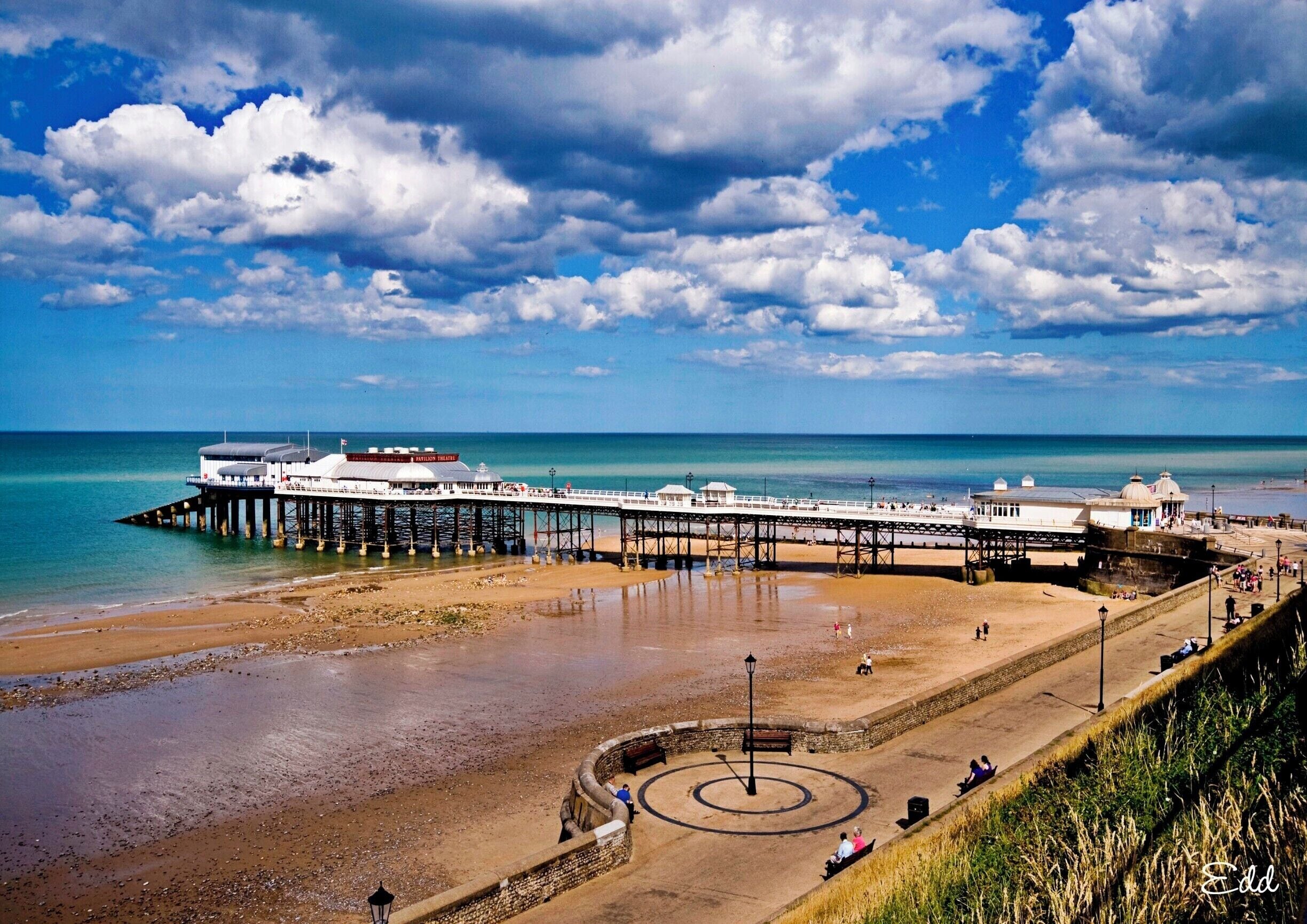 A pier extending into the sea with a beach and a circular monument in the foreground.