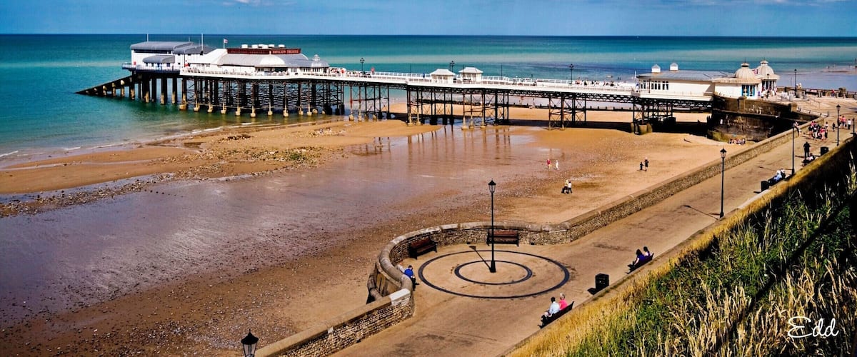 A pier extending into the sea with a beach and a circular monument in the foreground.