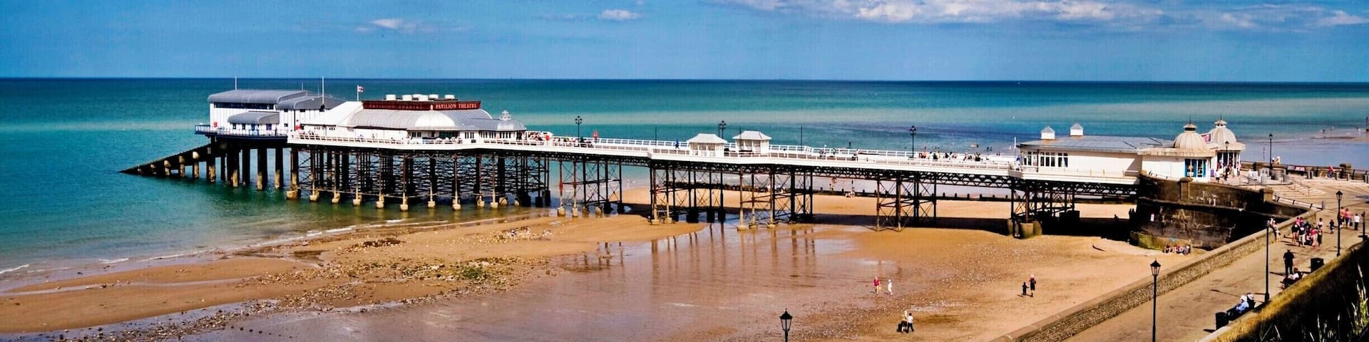 A pier extending into the sea with a beach and a circular monument in the foreground.