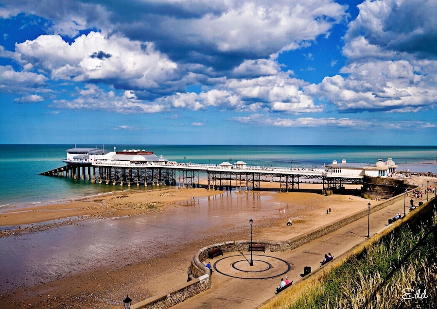 A pier extending into the sea with a beach and a circular monument in the foreground.