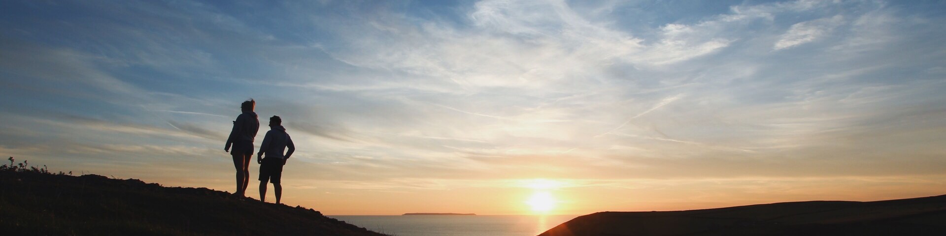 Two people silhouetted against a sunset on a grassy hill with a body of water in the distance.