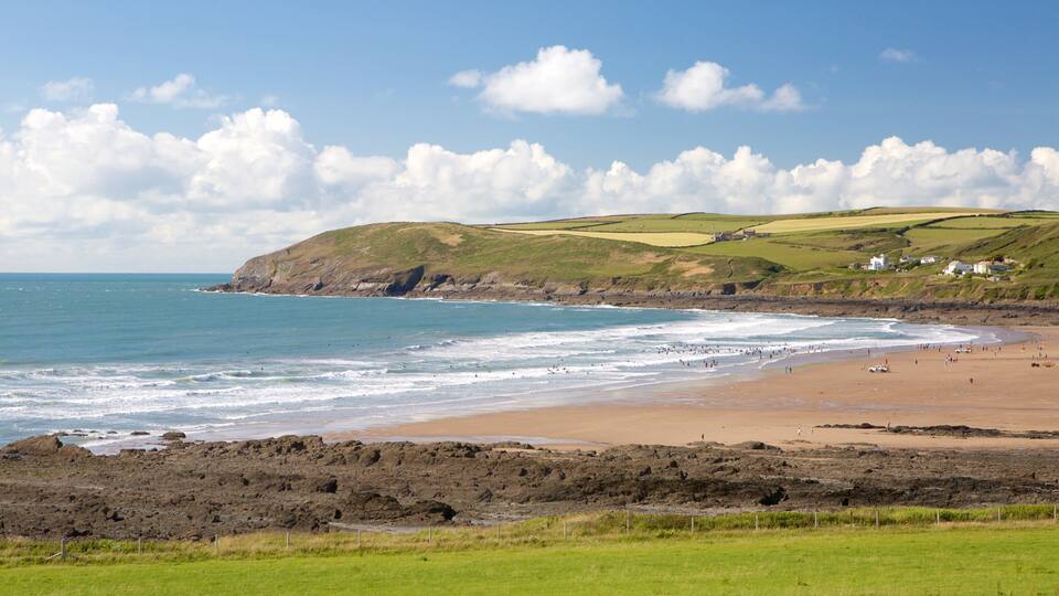 A coastal landscape with a sandy beach, rocky shoreline, and green hills in the background.