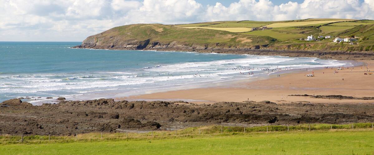 A coastal landscape with a sandy beach, rocky shoreline, and green hills in the background.