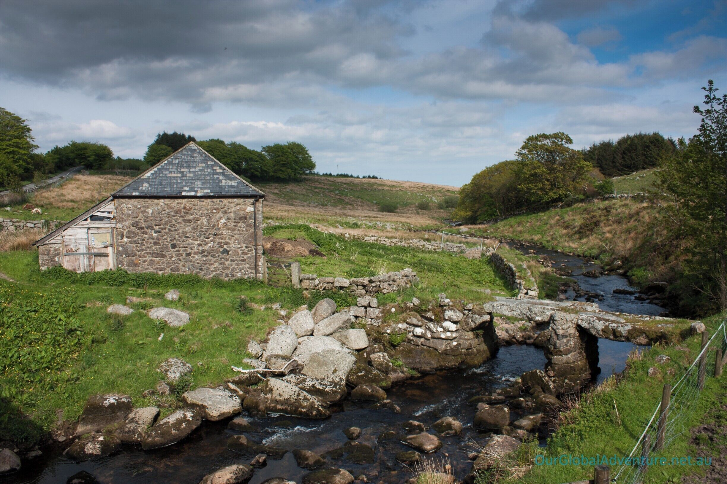 A stone house with a sloped roof, a stone bridge over a stream, and a grassy landscape with trees in the background.