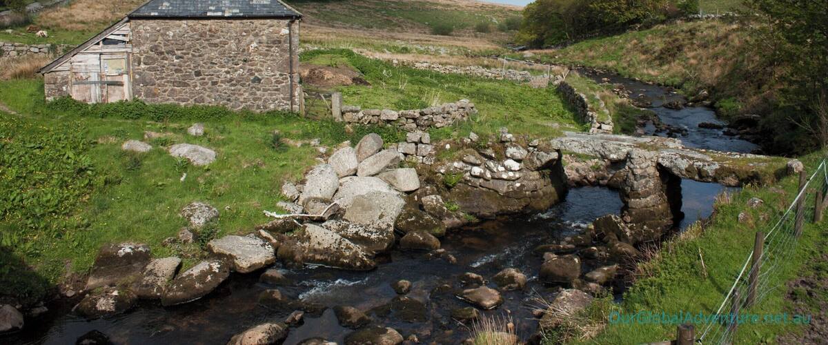 A stone house with a sloped roof, a stone bridge over a stream, and a grassy landscape with trees in the background.
