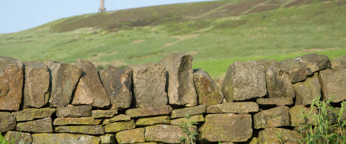 A stone wall in the foreground with a grassy hill and a lighthouse in the background.