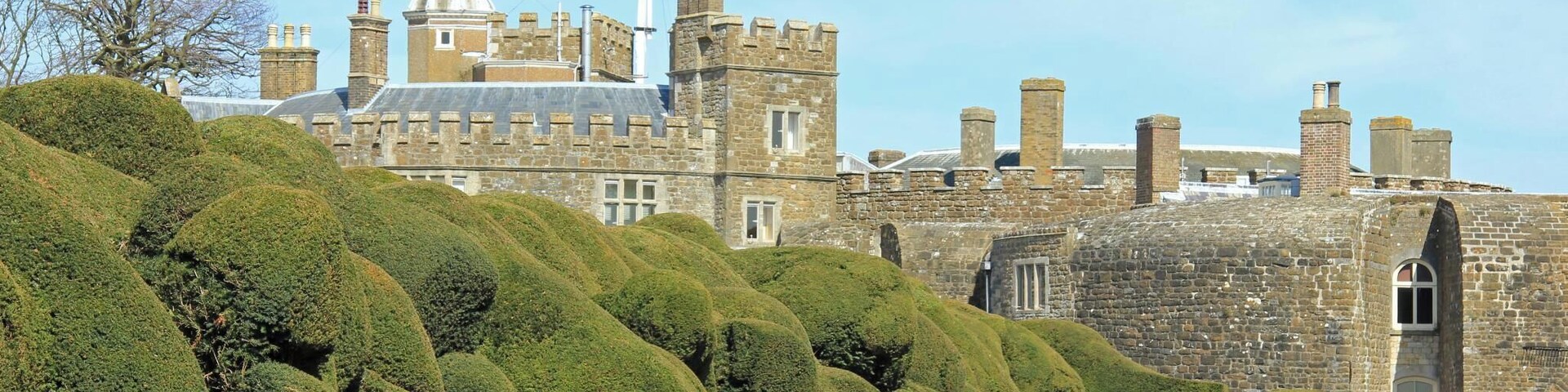 Un castillo de piedra histórico con una torre destacada, rodeado de setos cuidados y un cielo despejado.