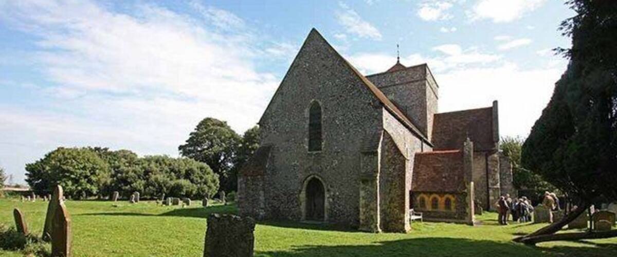 A stone church with a triangular roof and a bell tower, surrounded by a grassy field and trees.