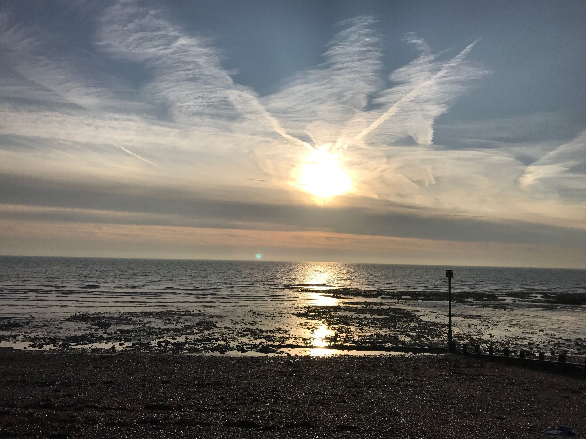 A sunset over a rocky shoreline with the sun partially obscured by clouds.