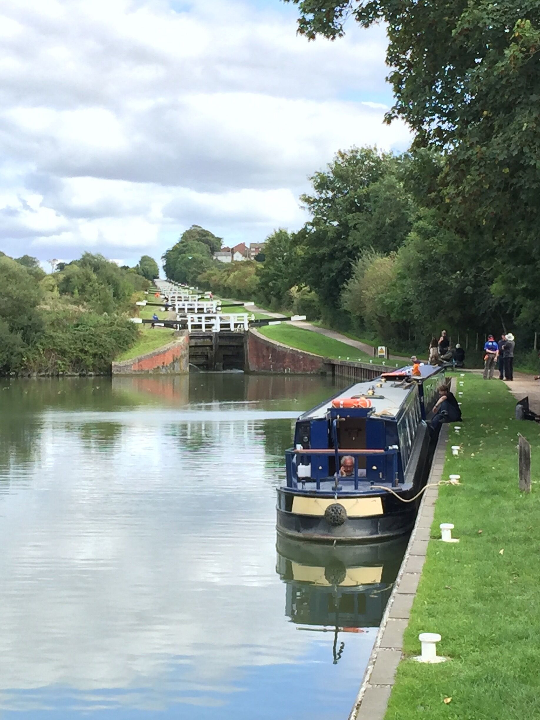 A narrowboat docked at a canal lock with a grassy bank and trees in the background.