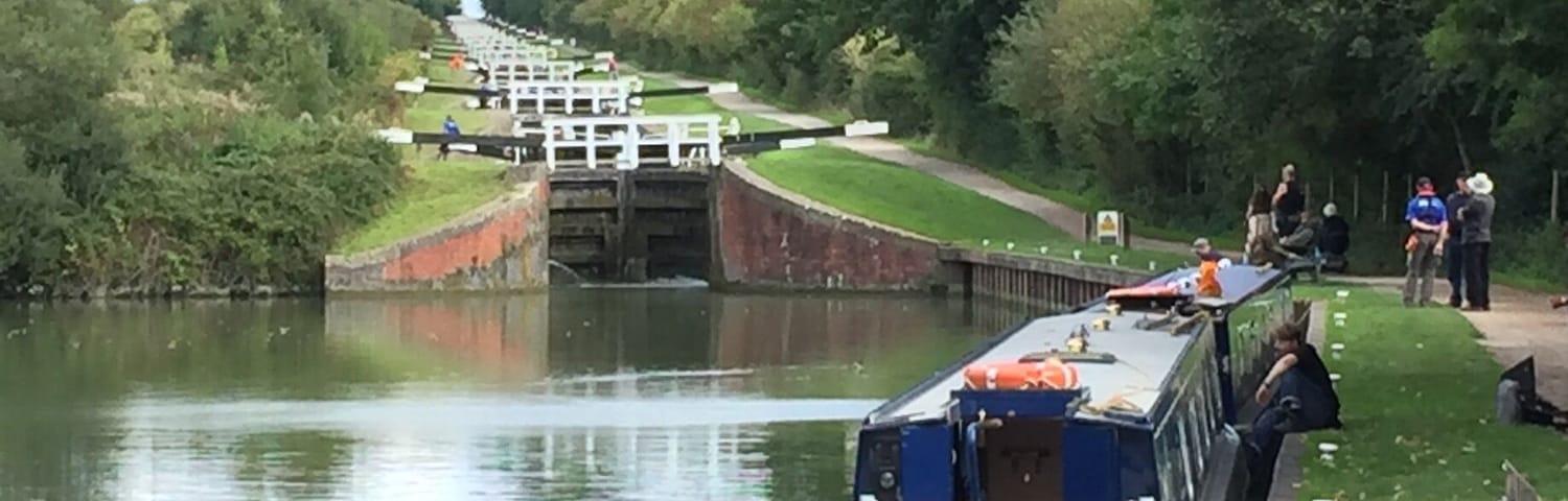 A narrowboat docked at a canal lock with a grassy bank and trees in the background.