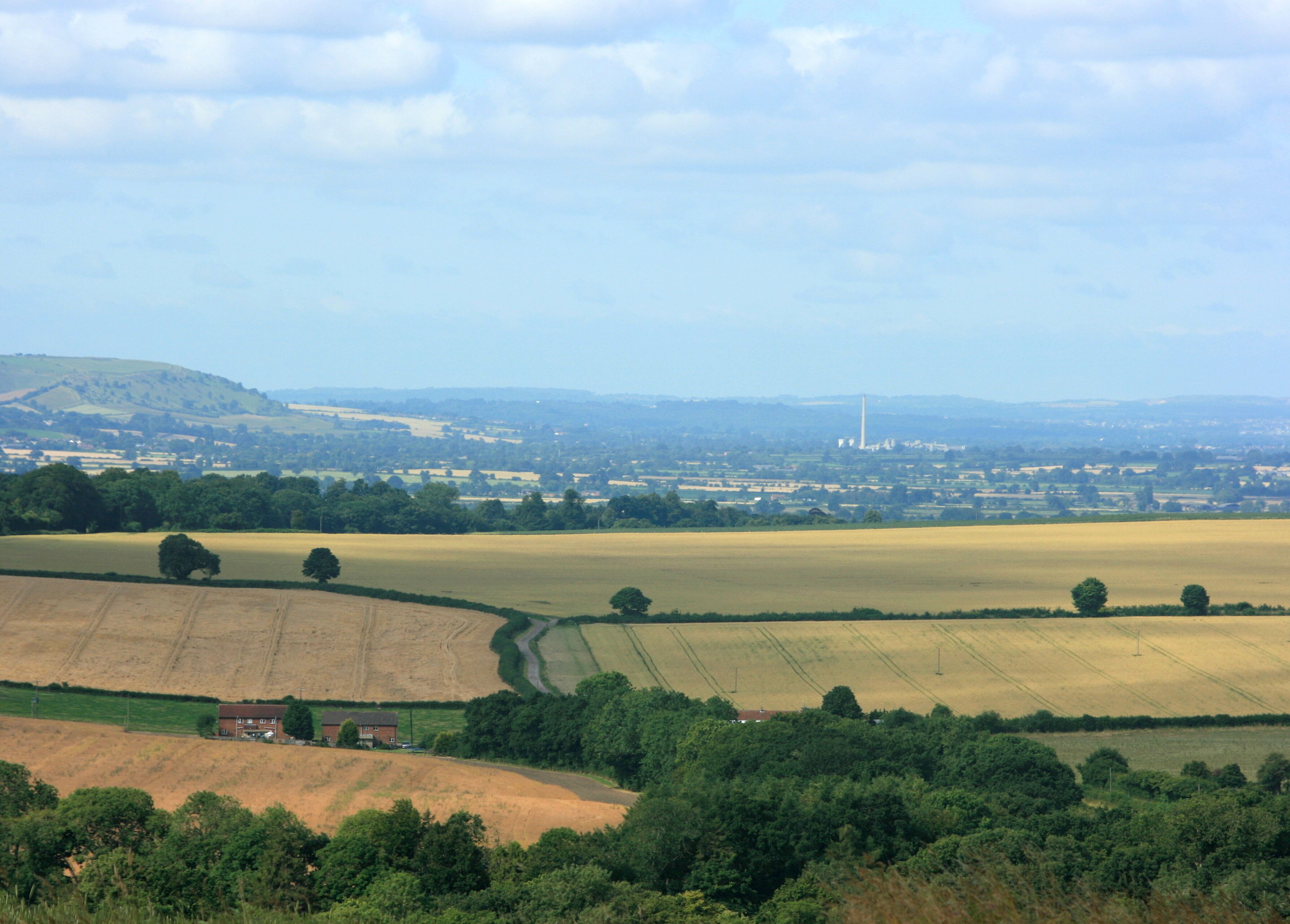 West from Etchilhampton Hill trigpoint (2). Swinging the camera north from 901094 brings the Lafarge Cement Works chimney ST8852 at Westbury into view. Bratton Camp is still visible this time on the far left. Ripening crops below the hill, mostly wheat, and farmland beyond complete the scene.