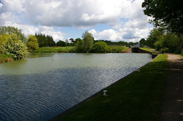 Une rivière paisible, bordée d’une rive herbeuse, avec des arbres et un pont en arrière-plan.