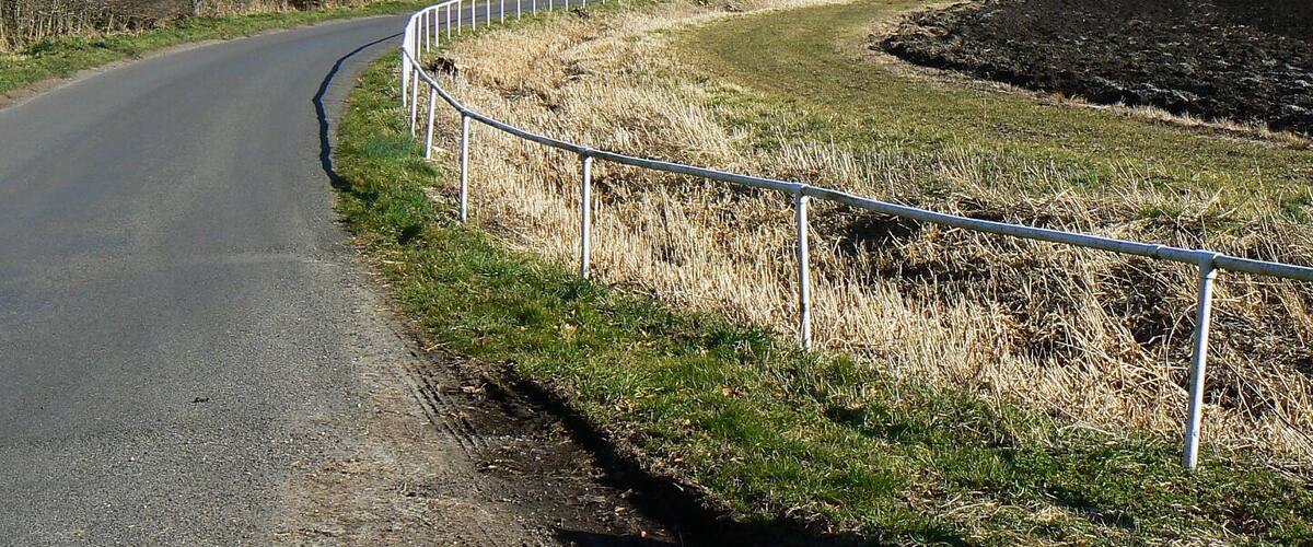 A rural road curving through fields with a metal guardrail, a building in the distance, and power lines overhead.