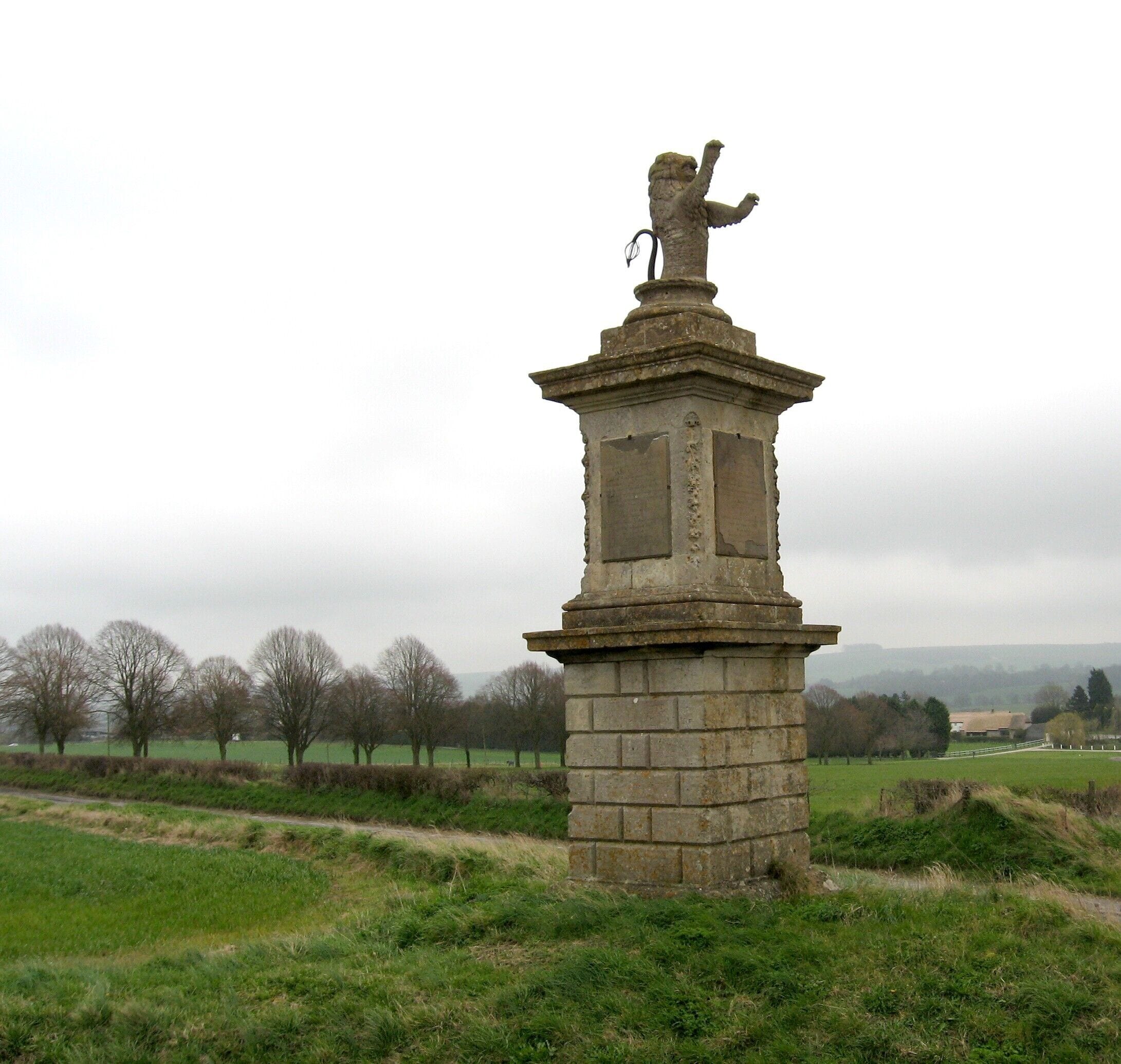 Un monumento de piedra con una estatua de león en la parte superior, rodeado de un paisaje rural con árboles y campos.