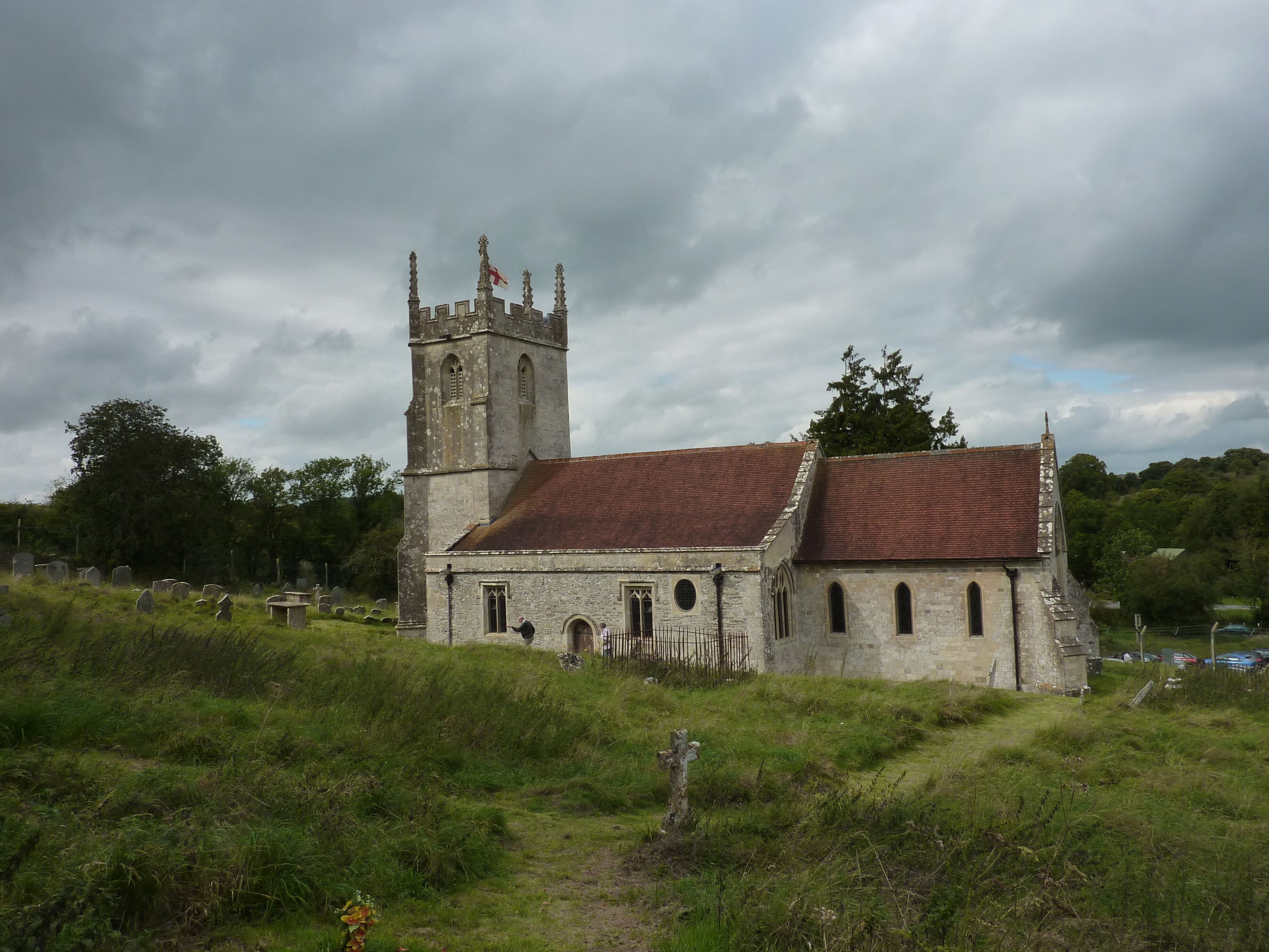 A historic church with a tall tower and a red-tiled roof, surrounded by a cemetery and lush greenery.