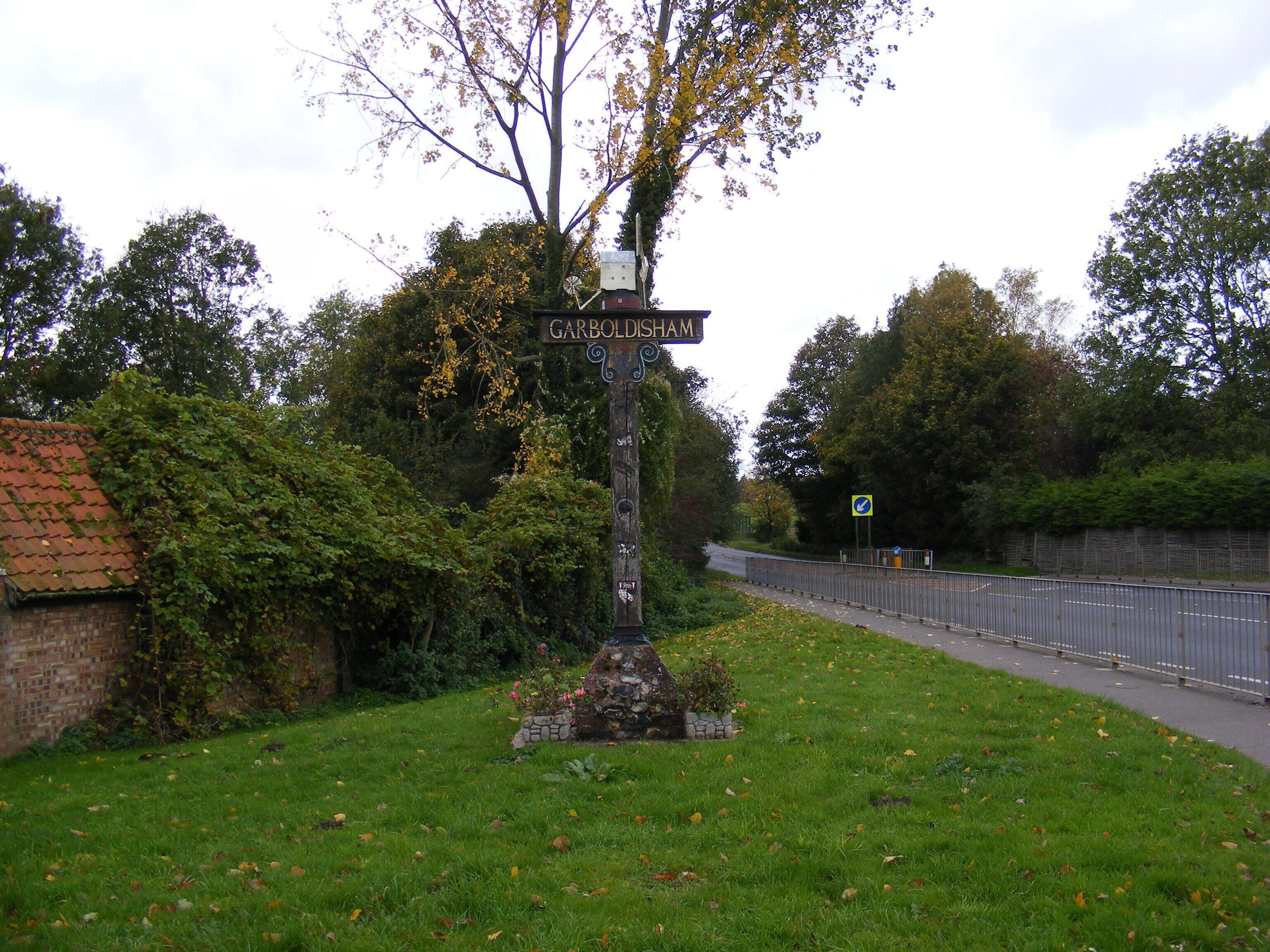 A signpost with 'GARBOLDISHAM' written on it, surrounded by greenery and a brick wall.