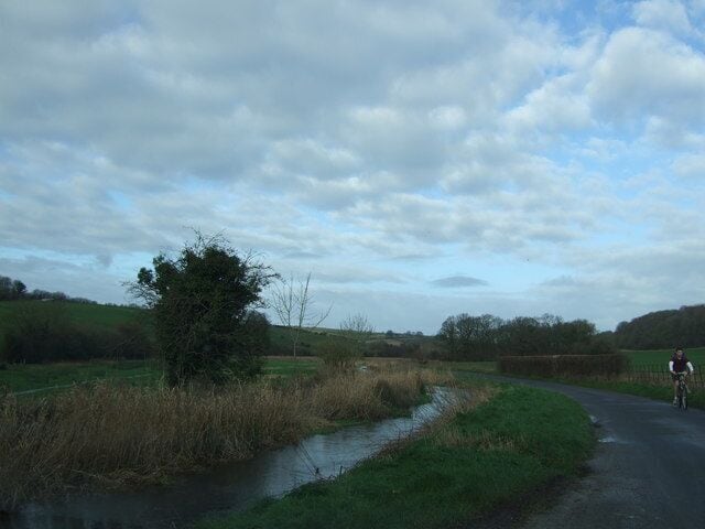 A rural road curving through a field with a small stream and a cyclist in the distance.