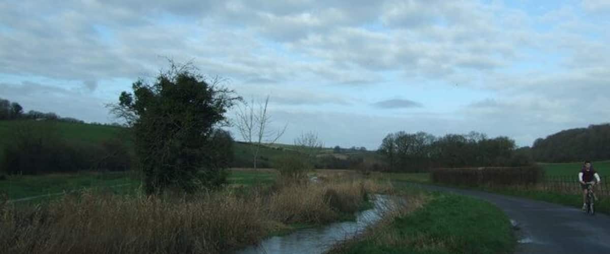 A rural road curving through a field with a small stream and a cyclist in the distance.