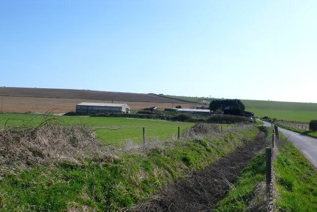 A rural landscape with a narrow road, green fields, and farm buildings.