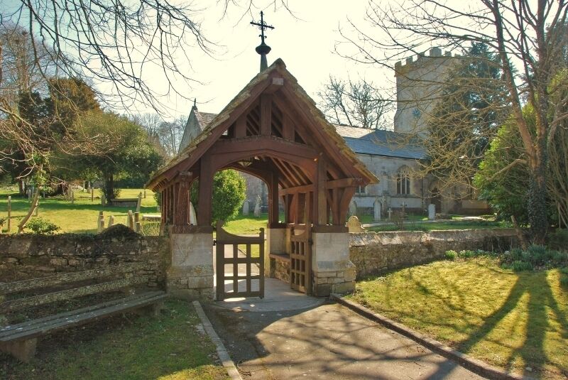 A wooden structure with a pointed roof and a cross at the top, surrounded by a stone wall and a grassy area with trees.