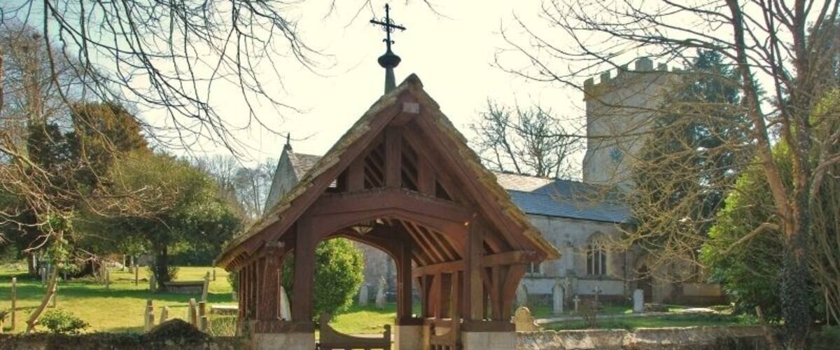 A wooden structure with a pointed roof and a cross at the top, surrounded by a stone wall and a grassy area with trees.