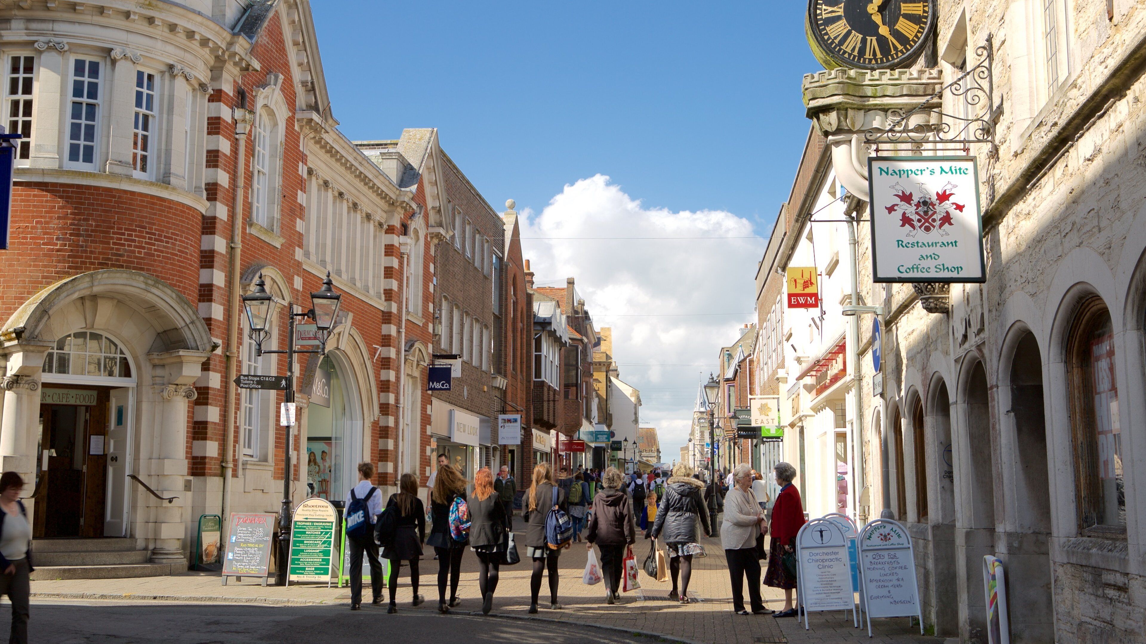 A bustling street with historic buildings, a clock tower, and various shops and cafes.