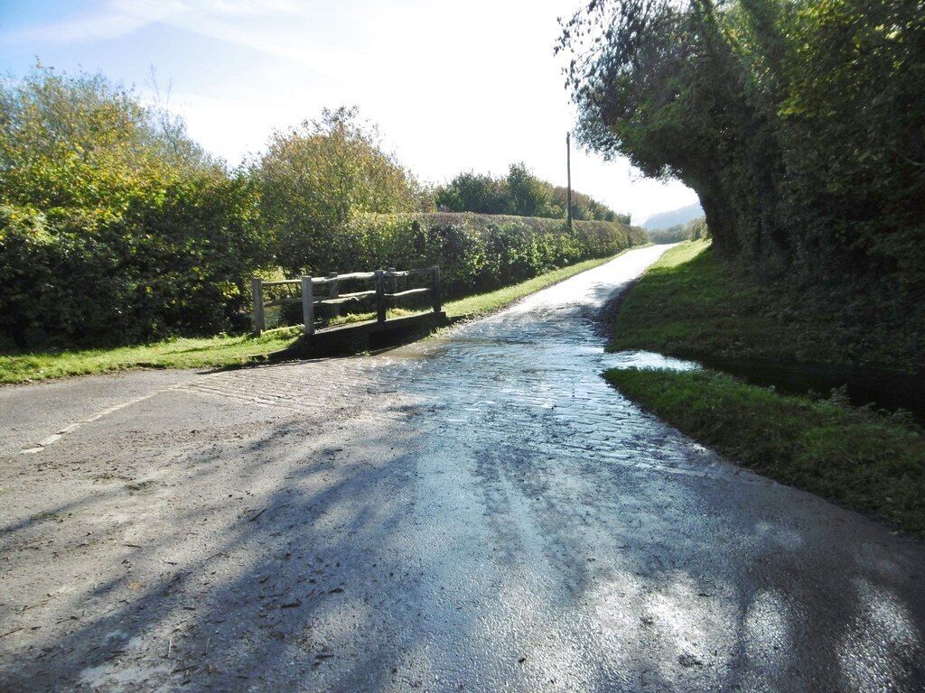 A narrow rural road with a wooden bridge, surrounded by lush greenery and a clear sky.