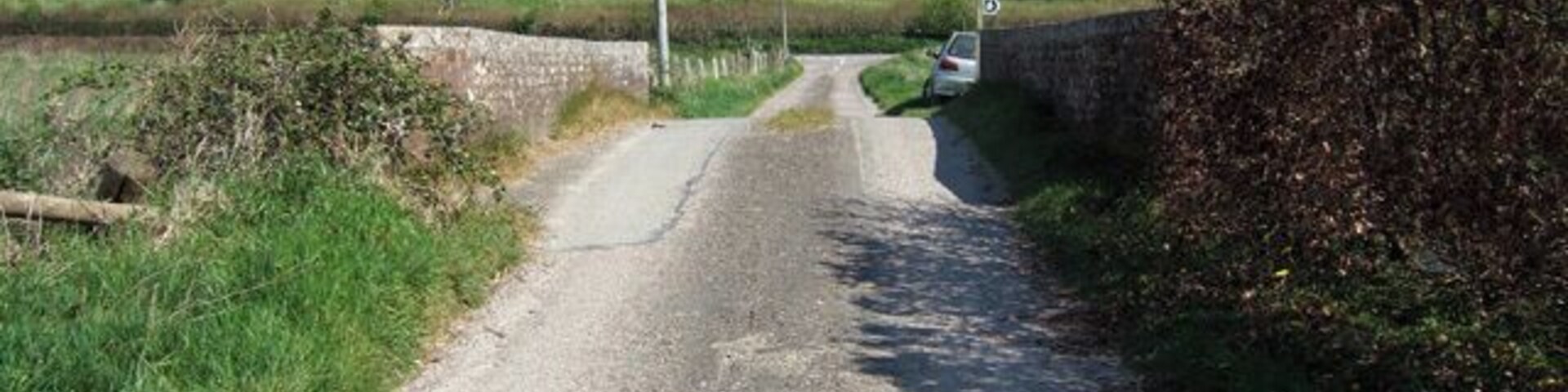 Uma estreita estrada rural ladeada por muros de pedra e sebes, serpenteando por uma paisagem verde com colinas ondulantes ao longe.