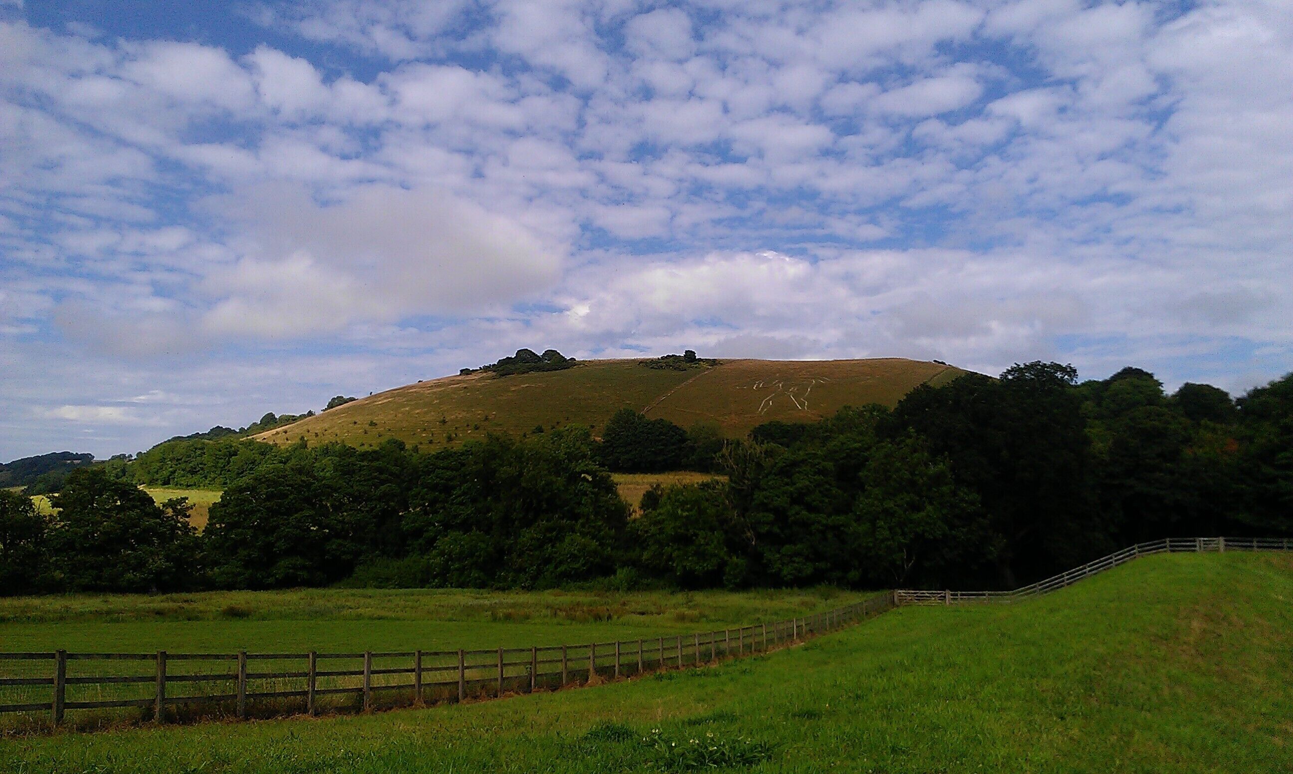 A grassy hill with a wooden fence in the foreground and a cloudy sky above.