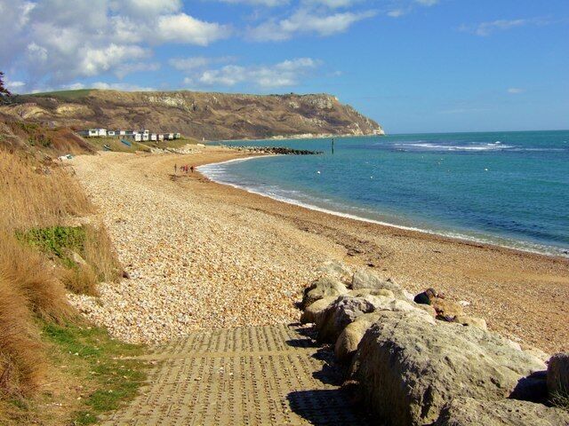 A pebble beach with a rocky shoreline, a clear blue sea, and a distant cliff.
