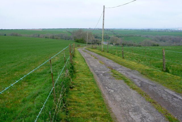 A rural road bordered by green fields and a wire fence.