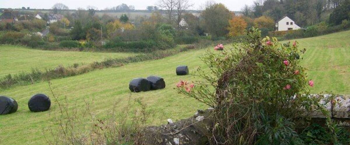 A rural landscape with hay bales, a stone wall, and a house in the background.