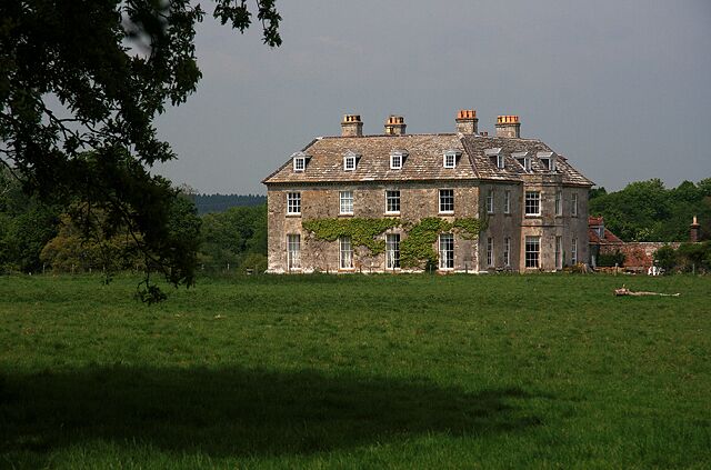 A large stone house with multiple chimneys, surrounded by a grassy field and trees.