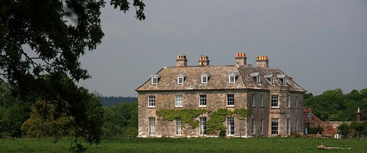 A large stone house with multiple chimneys, surrounded by a grassy field and trees.