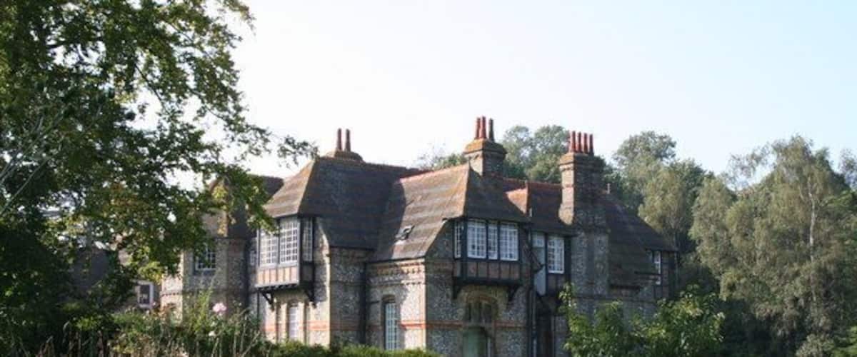 A large, historic stone house with a steeply pitched roof, surrounded by lush greenery and trees.