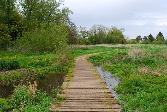 A wooden boardwalk along a water channel, surrounded by greenery and trees.